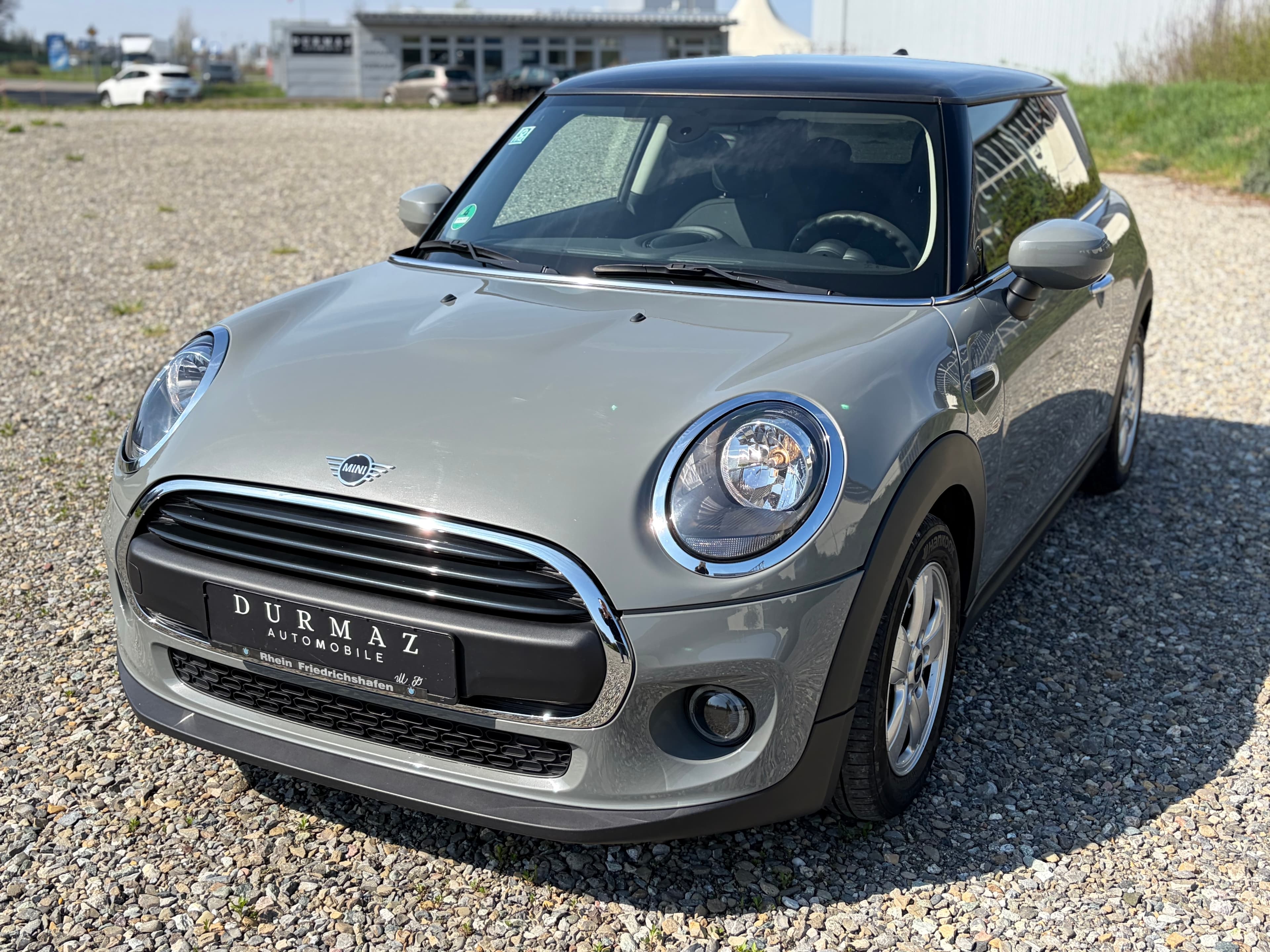 Grauer Mini Cooper mit schwarzem Dach parkt auf einem hellen Kiesplatz vor einem Autohaus.