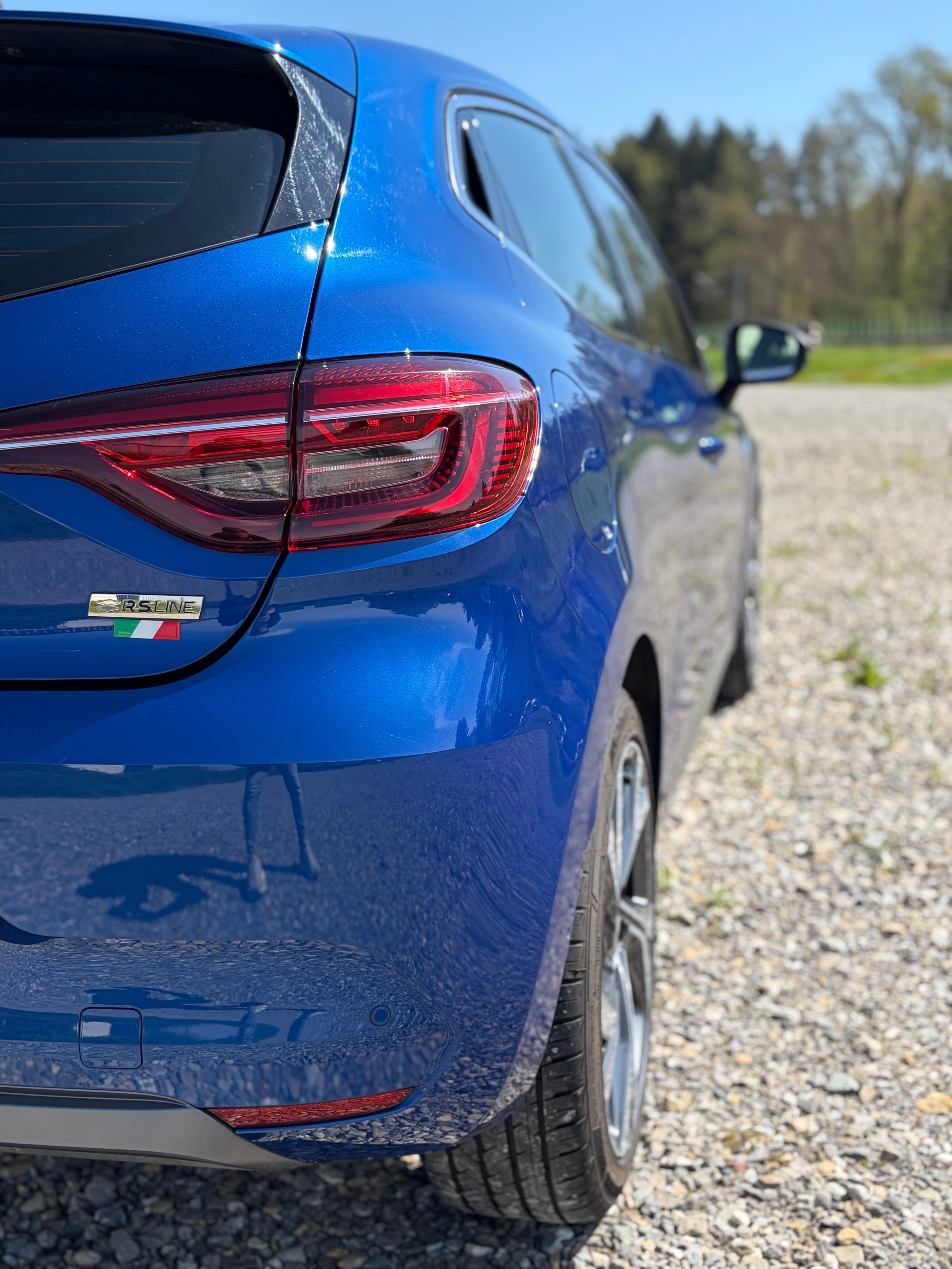 Nahaufnahme eines blauen Renault Clio RS Line mit markanten Rückleuchten auf einem sonnigen Schotterplatz.