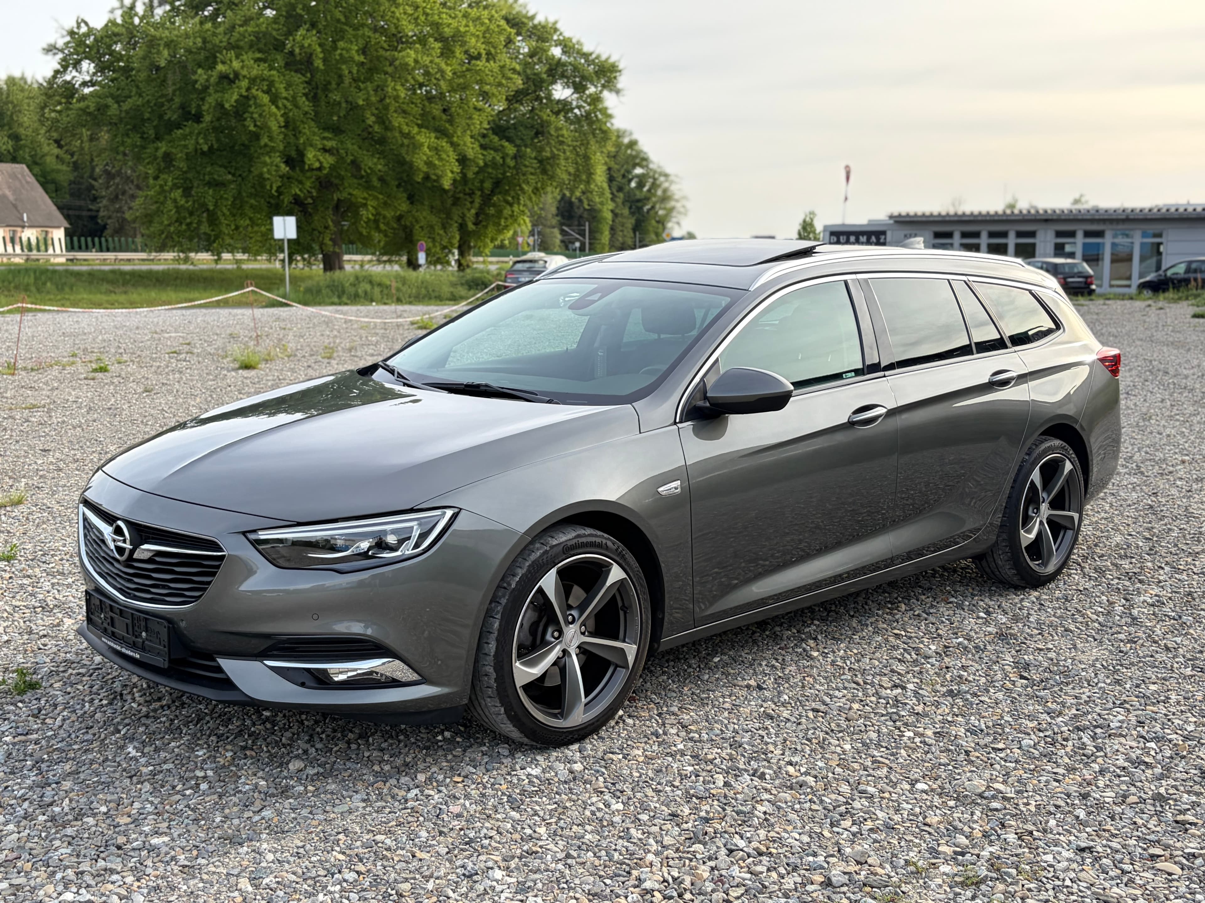 Grauer Opel Insignia Sports Tourer mit Alufelgen auf einem Kiesparkplatz vor grünen Bäumen.